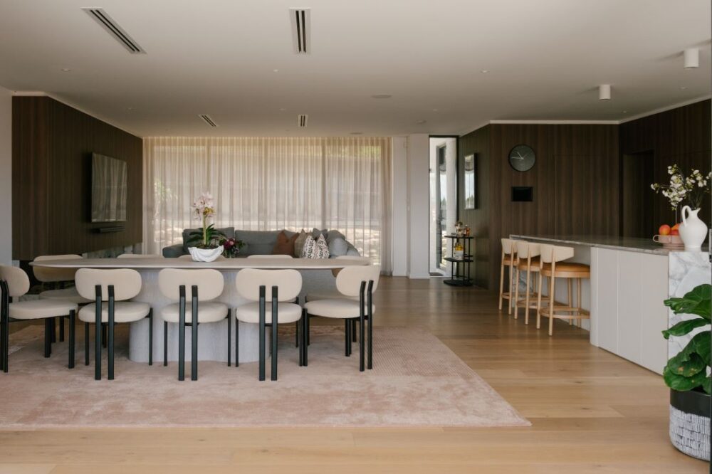A wide open plan living, dining, and kitchen area at Drummoyne House - Issy & H Creative. Features a modern oval dining table with cream minimalist chairs, integrated dark woodgrain wall panelling, a kitchen island with timber bar stools, and floor-to-ceiling sheer curtains in the background.
