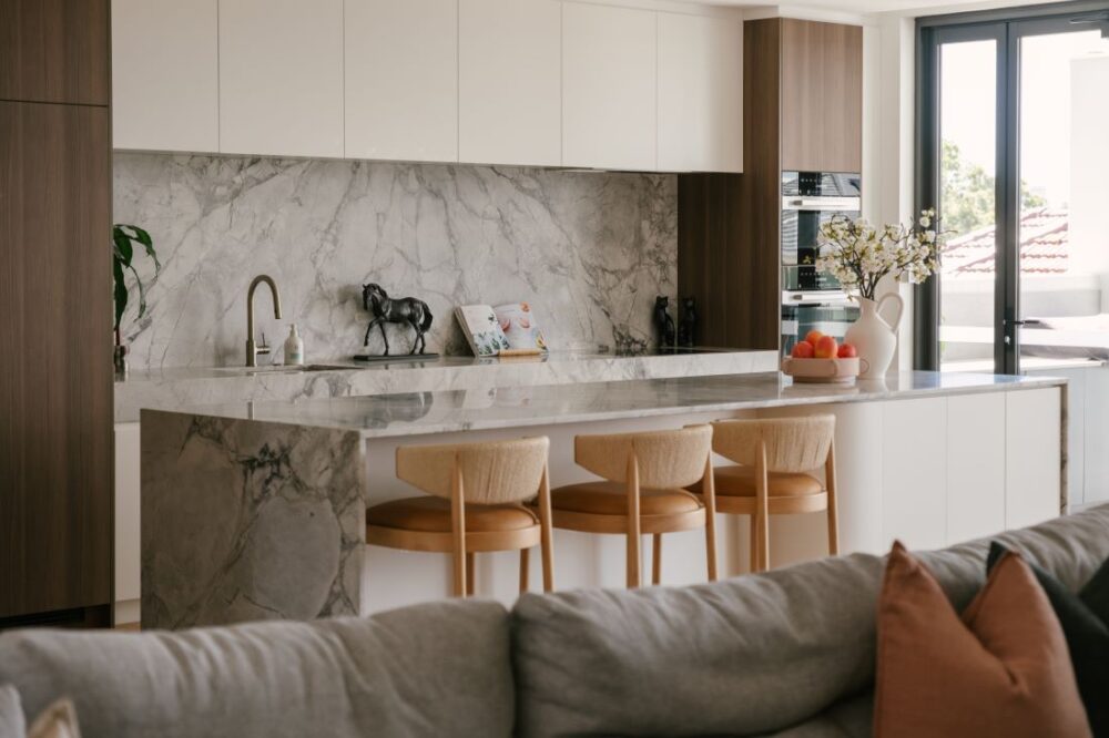 A view of the luxury kitchen at Drummoyne House - Issy & H Creative, taken from behind the living room sofa. Features a waterfall marble island bench with three woven-back timber bar stools, a matching stone splashback, white overhead cupboards, and integrated dark woodgrain pantry joinery.