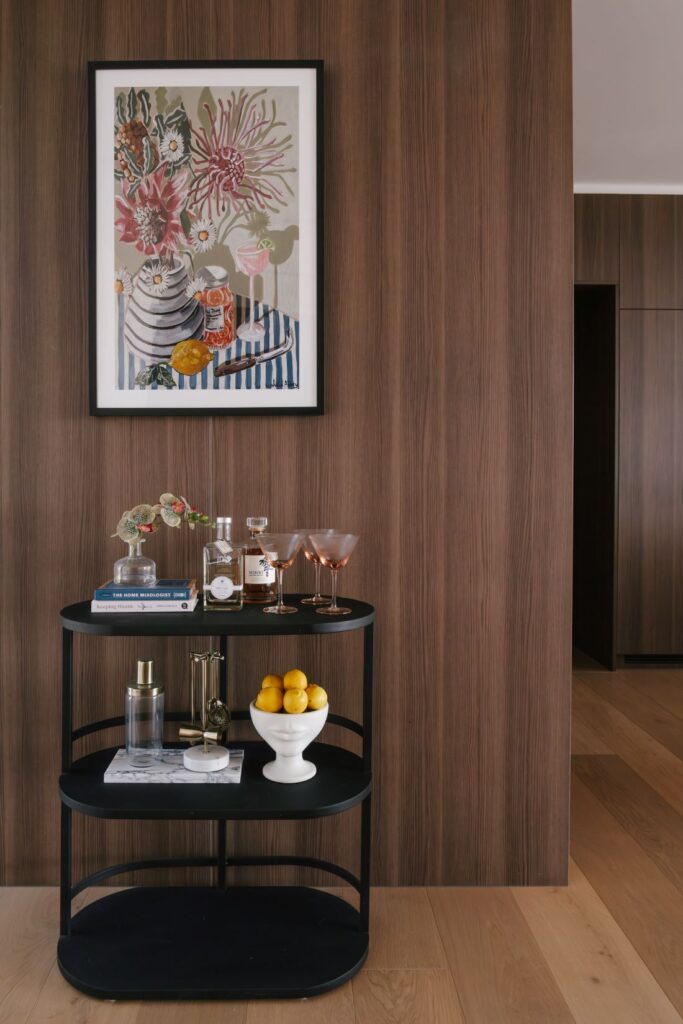 A beautifully styled three-tier black metal bar cart at Drummoyne House - Issy & H Creative. Set against a warm vertical timber panelling wall, featuring cocktail glassware, spirits, design books, a white ceramic face bowl filled with lemons, and a framed botanical still-life art piece hanging above.