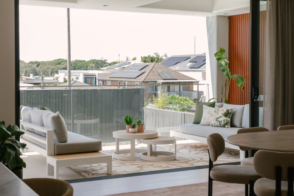 A wide-angle view of the modern coastal outdoor balcony at Drummoyne House - Issy & H Creative. Features two white-framed outdoor sofas with grey cushions, tropical accent pillows, nested round timber-topped coffee tables, a large patterned outdoor rug, and a warm vertical timber batten feature wall looking out over a glass balustrade.