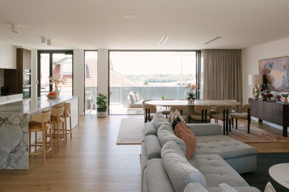 An expansive open plan living room, dining area, and kitchen at Drummoyne House - Issy & H Creative. Features a large grey modular sofa, a marble waterfall kitchen island with timber bar stools, an oval dining table, and large glass sliding doors that open up to a spacious alfresco balcony with outdoor seating.