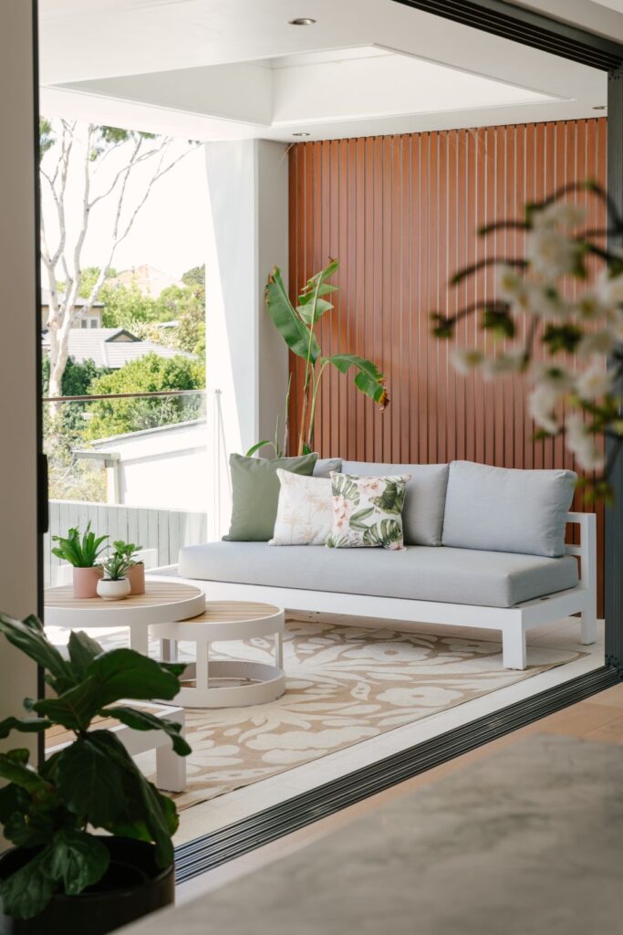 A modern coastal outdoor balcony at Drummoyne House - Issy & H Creative. Featuring a white-framed outdoor sofa with grey cushions, tropical print pillows, nested round timber coffee tables, and a warm vertical timber batten feature wall.
