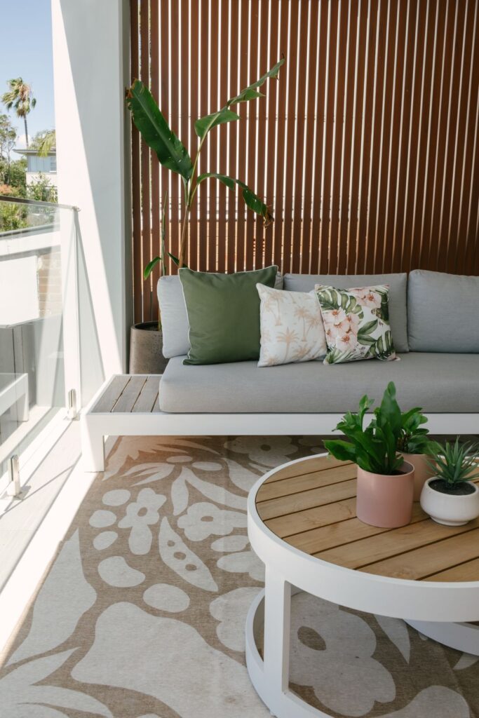 A styled close-up of an outdoor balcony area at Drummoyne House - Issy & H Creative. Features a white framed sofa with light grey cushions, tropical and olive green throw pillows, a large floral patterned outdoor rug, and a round timber-top coffee table with potted plants.