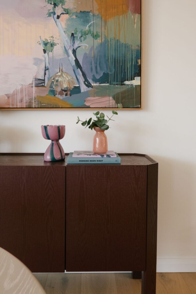 A beautifully curated interior styling vignette at Drummoyne House - Issy & H Creative. Features a dark stained timber sideboard cabinet topped with design books, a terracotta vase with eucalyptus branches, an eclectic green and pink checkered goblet vessel, and a large abstract landscape art piece hanging on the wall.