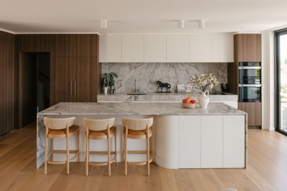 A luxury open plan kitchen design at Drummoyne House - Issy & H Creative. Features a large marble island bench with a curved profile, three light timber bar stools with woven backs, integrated dark woodgrain cabinetry, a matching marble splashback, and premium built-in double ovens.