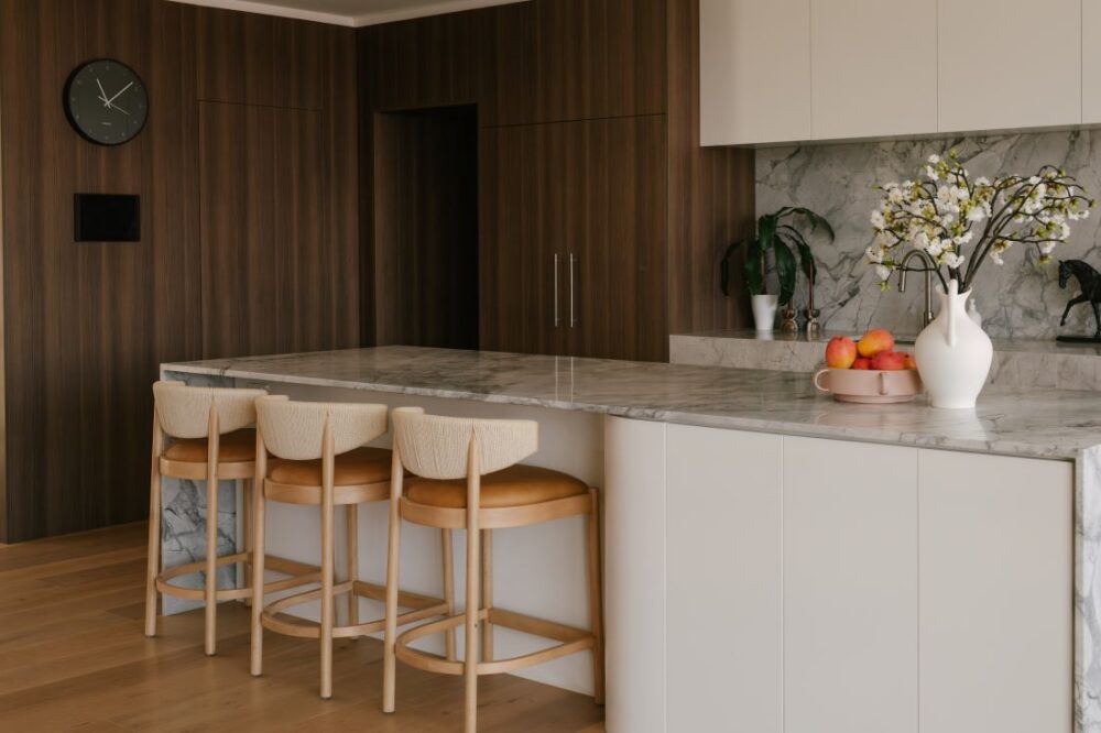A close-up view of the kitchen island bench at Drummoyne House - Issy & H Creative. Highlights the marble benchtops, three woven-back timber bar stools, a curved white island base, and rich dark woodgrain wall panelling with an integrated hidden door and black wall clock.