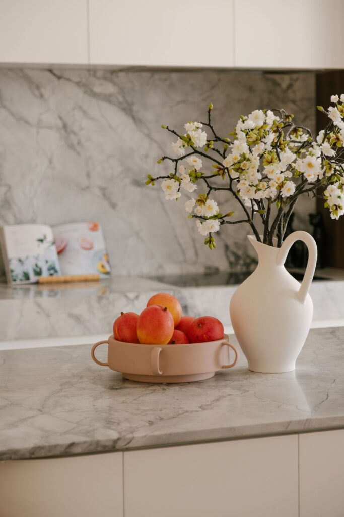 A close-up kitchen styling vignette at Drummoyne House - Issy & H Creative. Set on a grey veined marble benchtop, featuring a matte white ceramic pitcher vase with white cherry blossom branches, and a blush pink terracotta fruit bowl filled with fresh mangoes.