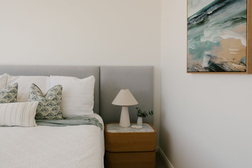 A serene coastal bedroom design at Drummoyne House - Issy & H Creative. Features a bed with an extended light grey upholstered bedhead, patterned sage green and white pillows, an oak timber bedside table with a marble top, a minimalist cone table lamp, and an abstract ocean landscape art piece.