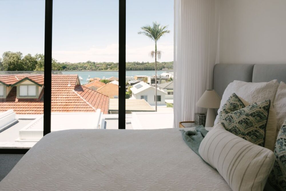 A luxury master bedroom at Drummoyne House - Issy & H Creative looking out to water views through large floor-to-ceiling windows. Features a bed dressed in white textured linen, patterned sage green accent pillows, a minimal bedside lamp, and a view of rooftops, a palm tree, and the river.