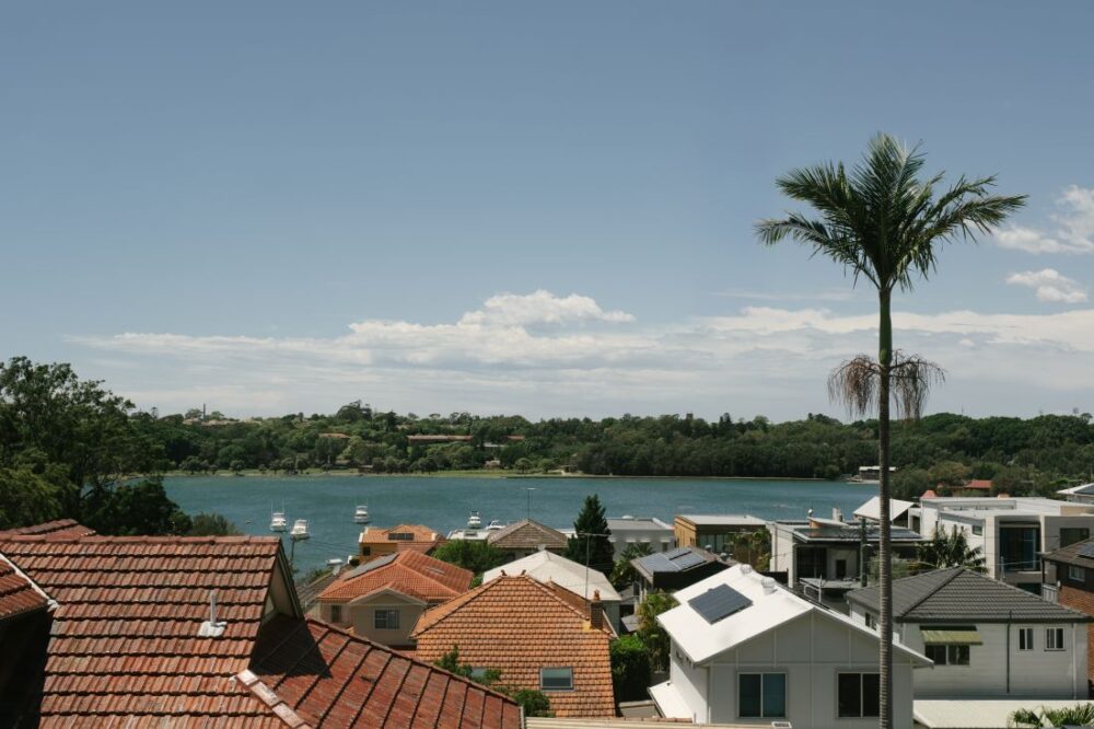 A panoramic outdoor view captured from Drummoyne House - Issy & H Creative. Showcases a scenic river or bay with moored boats, surrounded by suburban terracotta and modern white rooftops, green hillsides in the distance, and a tall palm tree under a clear blue sky.