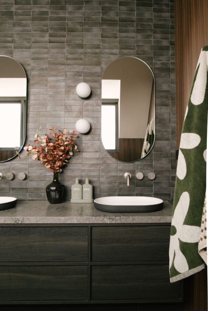 A luxury bathroom design at Drummoyne House - Issy & H Creative. Features a floating dark charcoal timber vanity with a grey stone top, a grey-rimmed basin with minimalist wall-mounted tapware, an oval mirror, textured grey subway wall tiles, and two round frosted globe wall sconces.