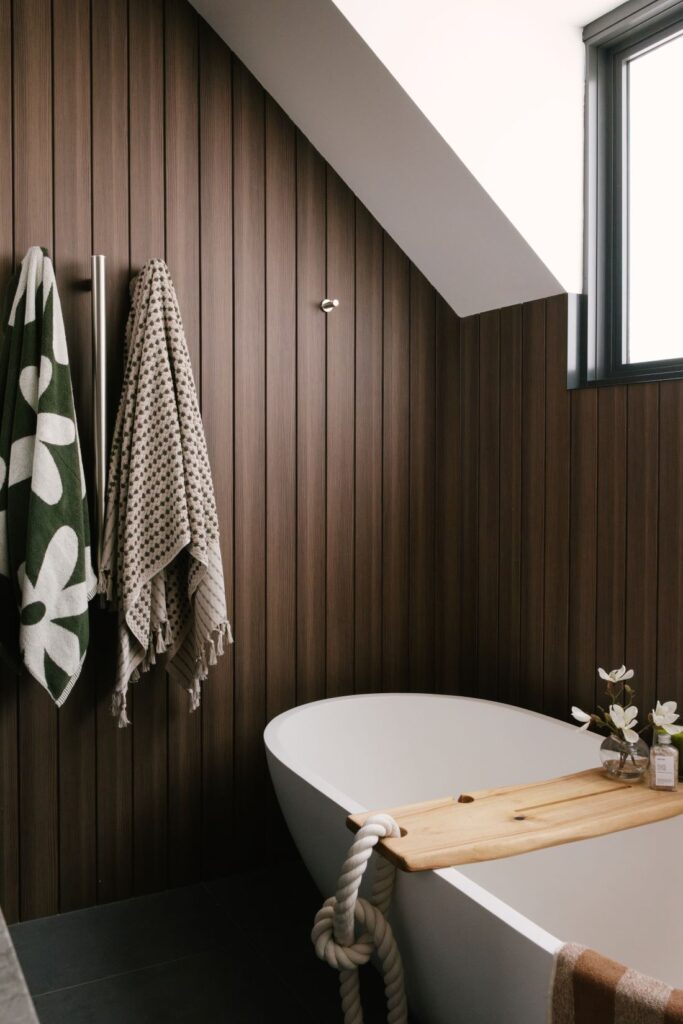 A luxury bathroom sanctuary at Drummoyne House - Issy & H Creative. Features a white freestanding oval bathtub tucked under a sloped ceiling, set against dark vertical timber wall panelling. A natural wood bath caddy with a rope detail holds a small vase of flowers next to hanging patterned towels.