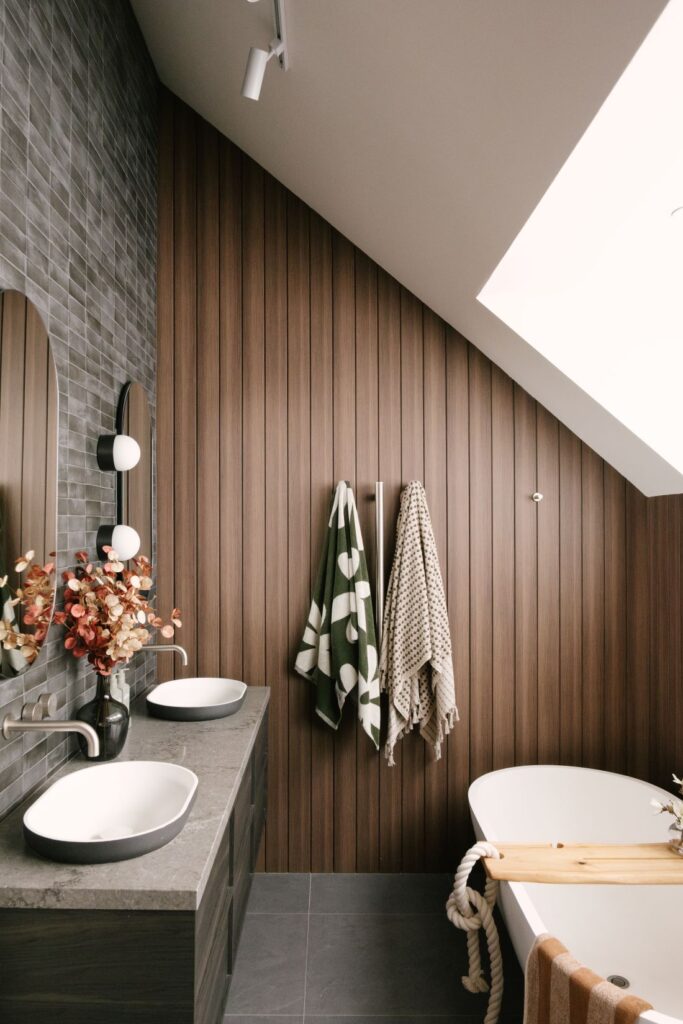 A full view of the luxury master ensuite bathroom at Drummoyne House - Issy & H Creative. Features a long double vanity with stone benchtops, grey tiled walls, oval mirrors, and a freestanding white bathtub situated under a sloped ceiling lined with dark vertical timber cladding.