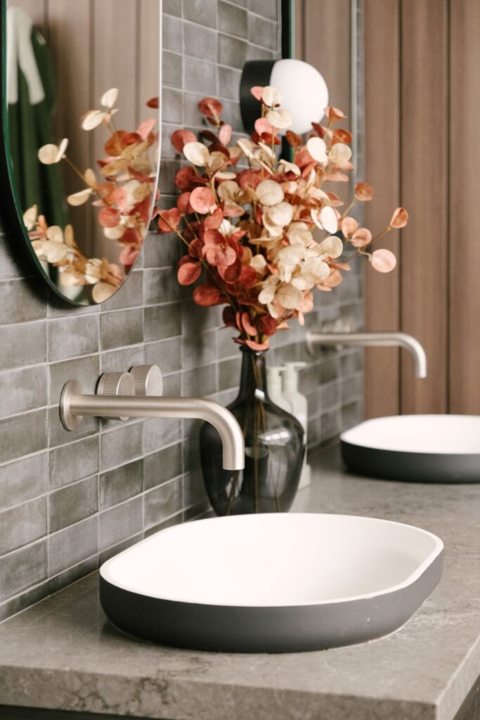 A close-up view of a luxury bathroom vanity setup at Drummoyne House - Issy & H Creative. Features a modern grey-rimmed countertop basin, sleek wall-mounted brushed nickel tapware, small grey subway wall tiles, a glass vase with dried eucalyptus branches, and a partial view of an oval mirror.
