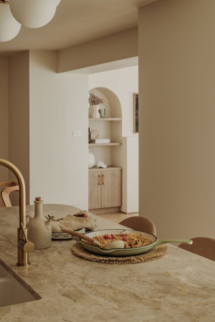 Marble kitchen island with brass faucet, skillet of pasta, ceramic oil bottle, neutral décor, and arched built-in shelving in background.
