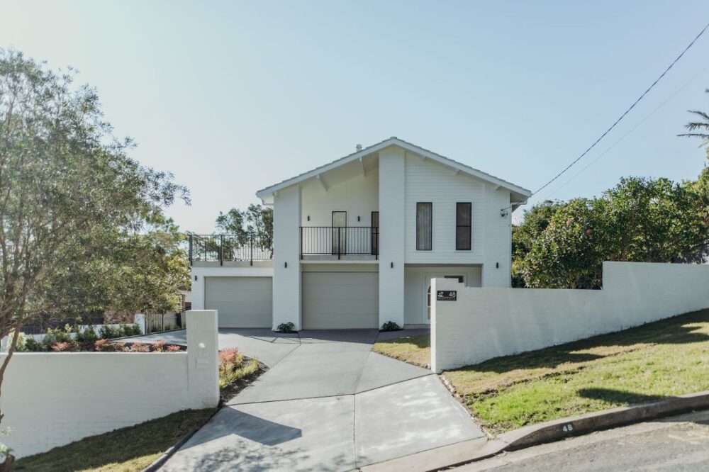 Modern two-story white house with triple garage, balcony, front driveway, landscaped garden, and sloping lawn.