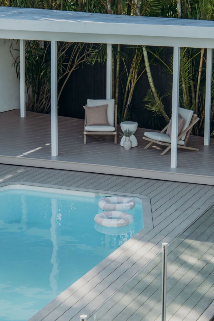 Outdoor pool deck with two lounge chairs, neutral cushions, small white side table, tropical greenery, and pool float in blue water.