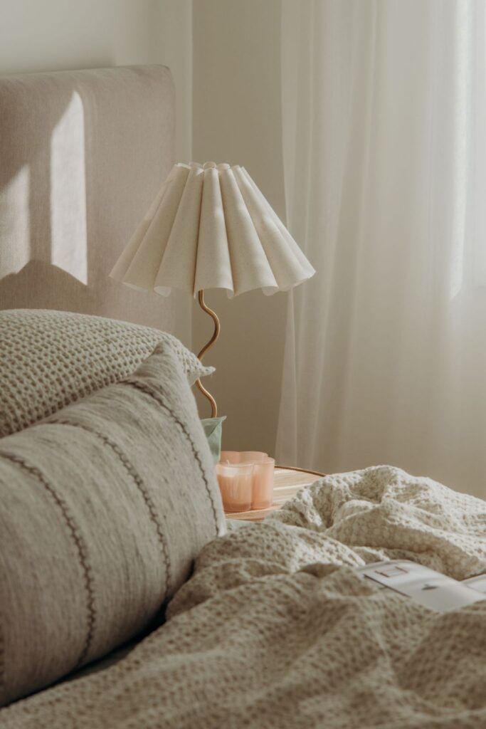 Bed with textured linen bedding, pleated table lamp on bedside table, soft natural light, and neutral bedroom decor.
