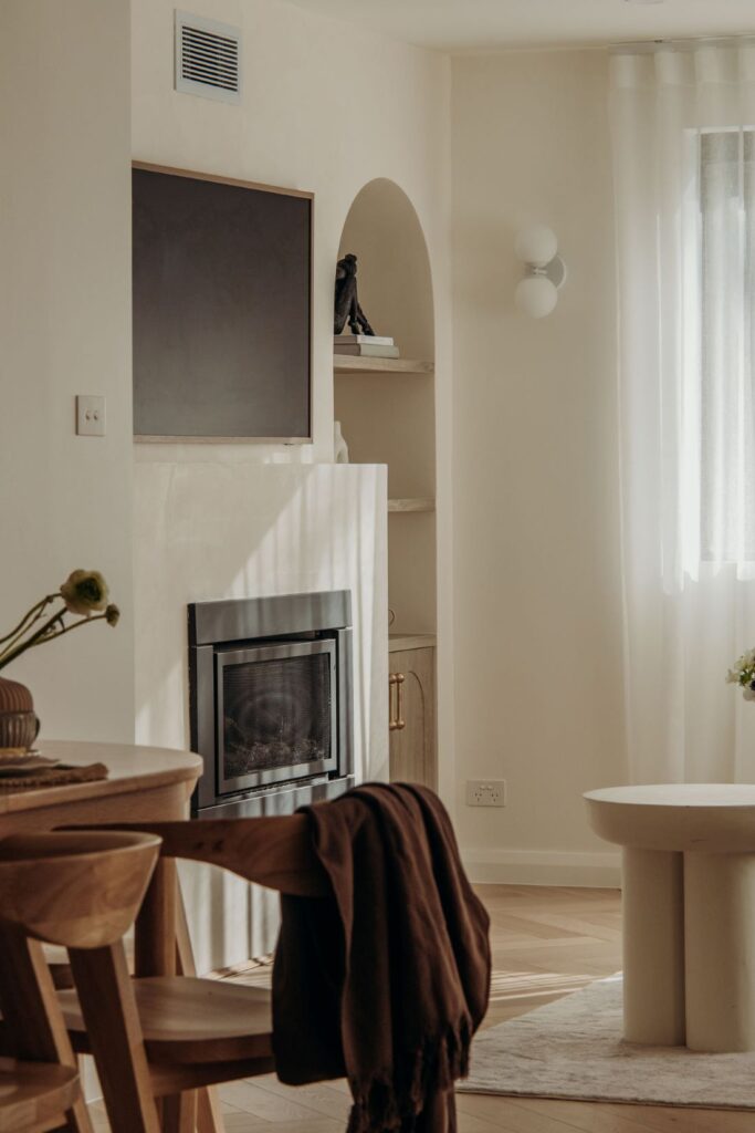 Minimalist living room with modern fireplace, built-in arched shelving, wall-mounted TV, timber dining chair, and soft natural light.