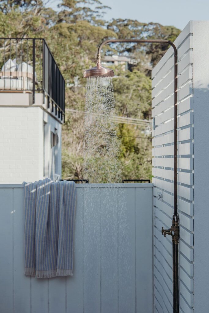 Copper outdoor shower with water flowing, white slatted privacy screen, striped towel, timber decking, and surrounding greenery.