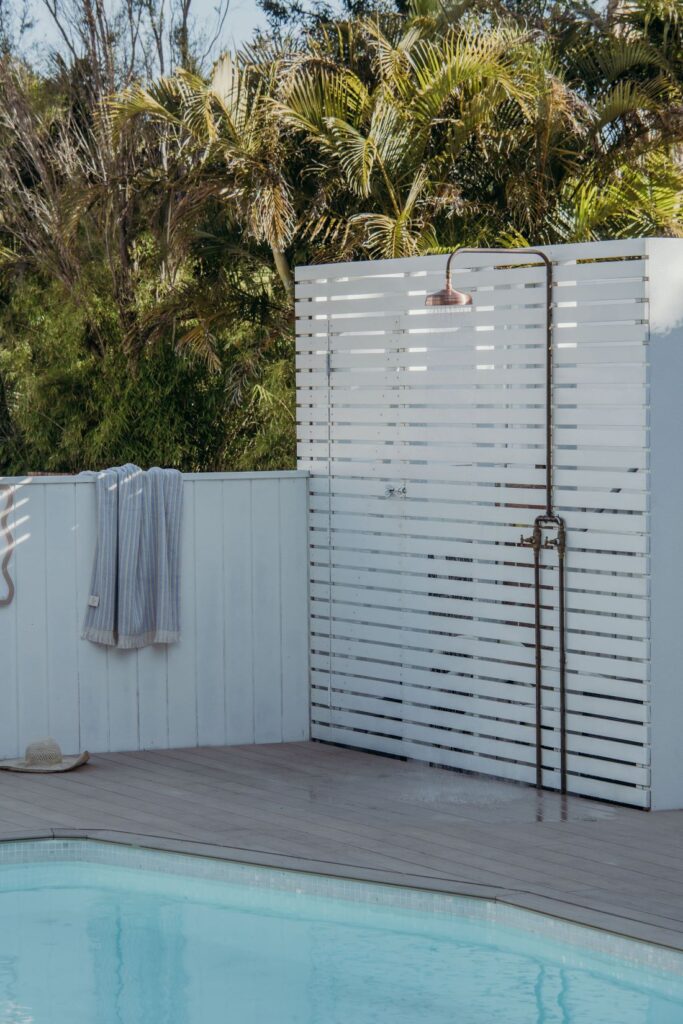 Outdoor poolside shower with white slatted screen, copper fixture, timber deck, striped towel, straw hat, and tropical greenery.