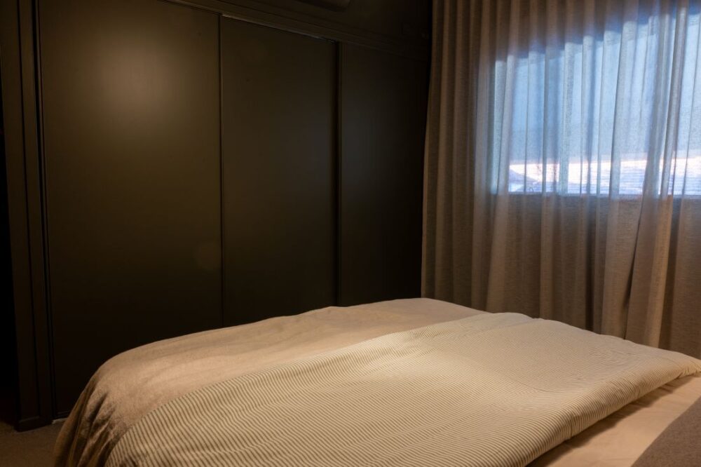 Bed with neutral bedding in bedroom with dark green wardrobe and sheer curtains