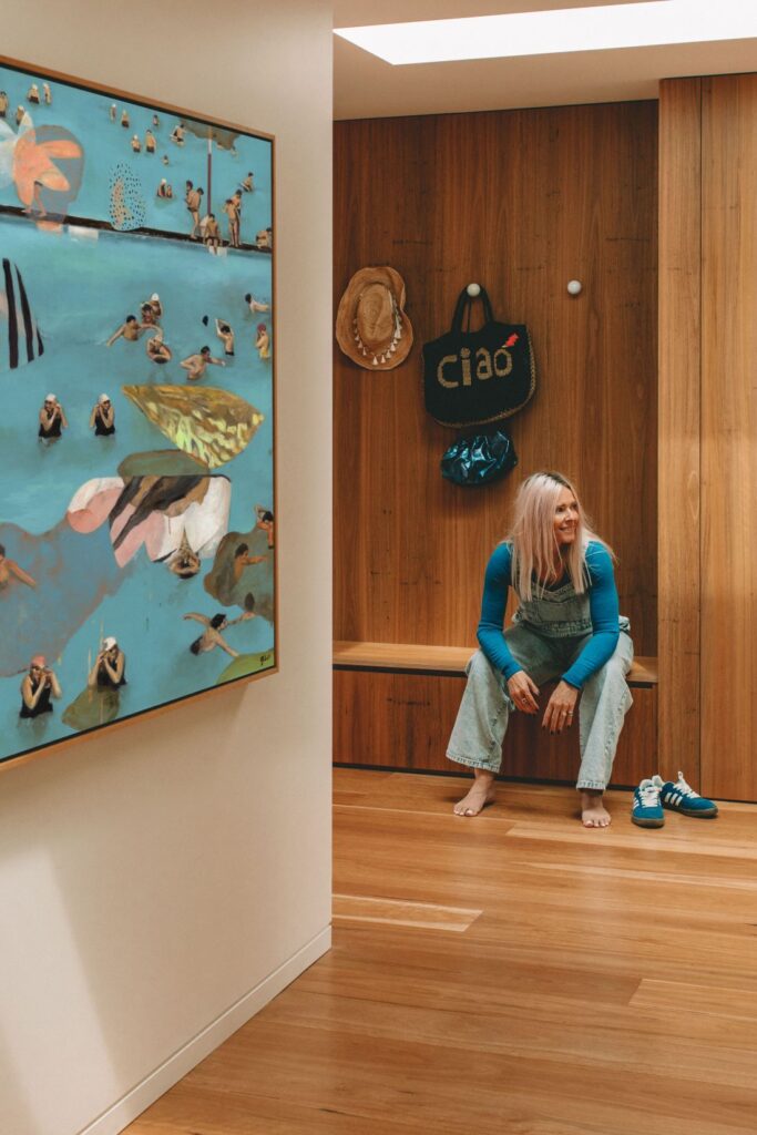 Artist sitting on bench beside large blue abstract figurative painting in hallway