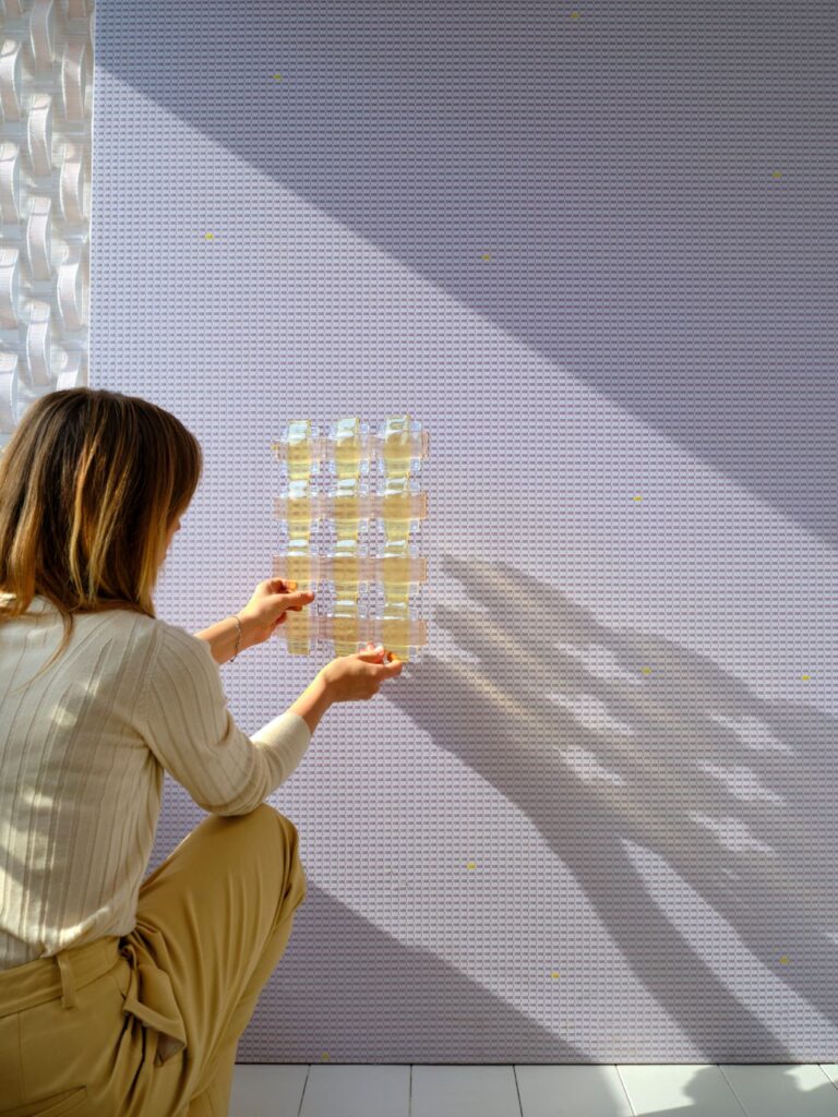 Designer holding a translucent grid-like material sample against a lavender textured wall in sunlight