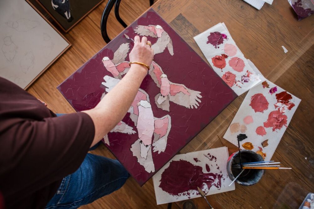 Overhead view of an artist applying thick paint to stylised pink and cream galahs on a burgundy canvas