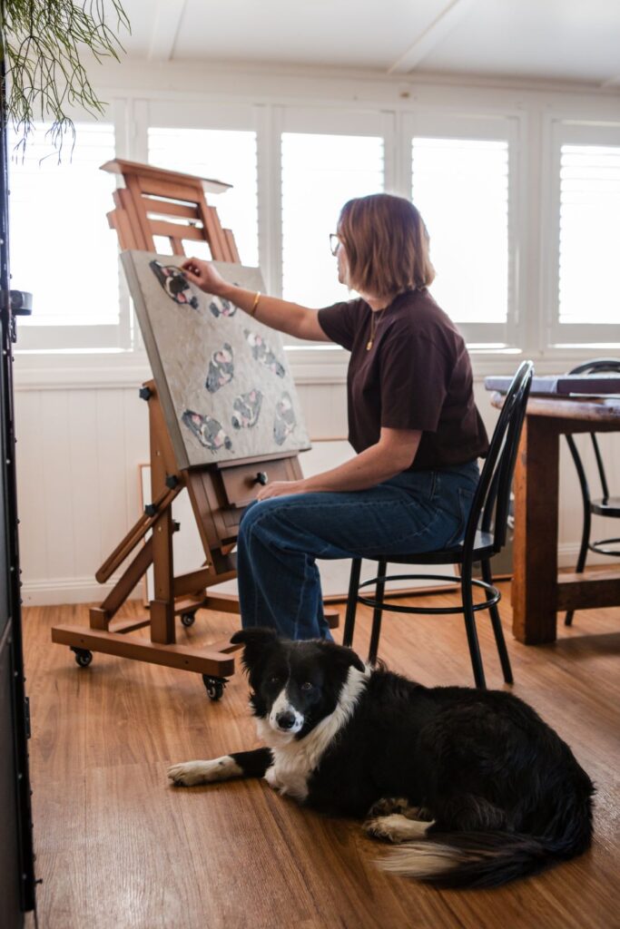 Artist Tanya Boyes working on a textured bird painting at an easel while her black and white border collie rests nearby