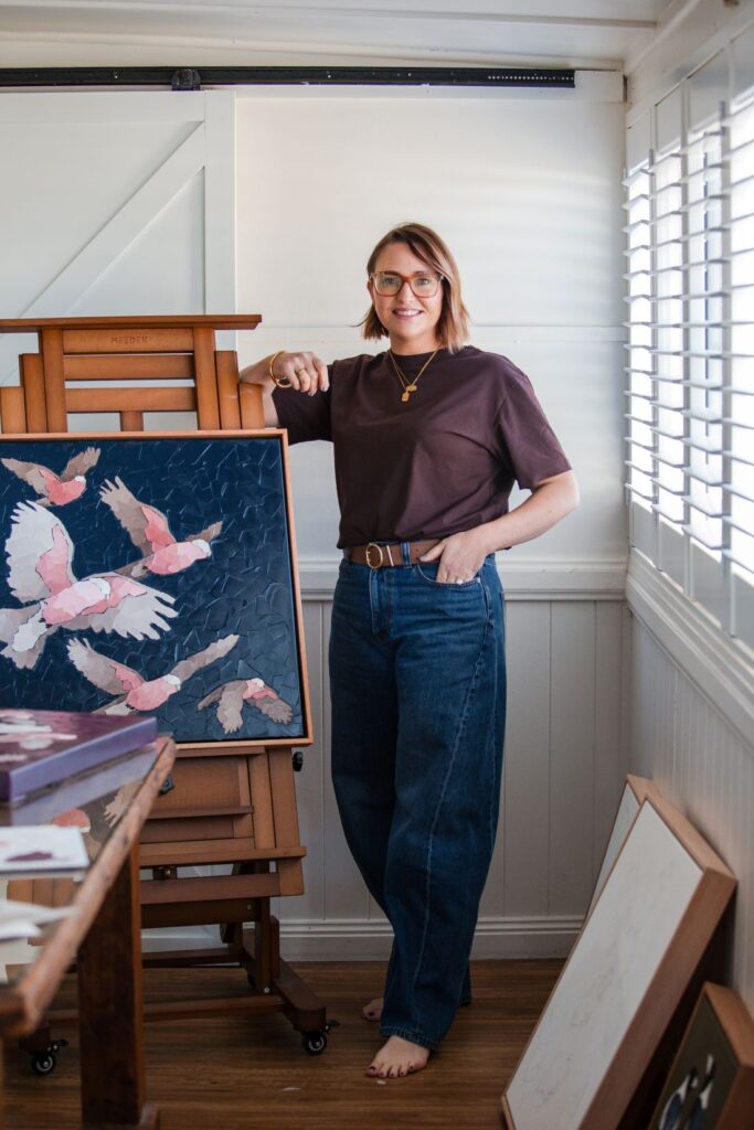 Artist Tanya Boyes standing beside a textured painting of pink galahs displayed on an easel in her Dalby home studio