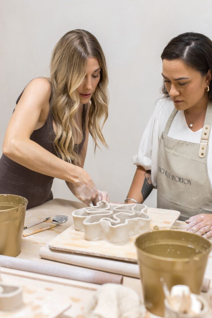 Ceramic instructor demonstrating hand-building techniques beside a workshop participant shaping an organic clay sculpture.