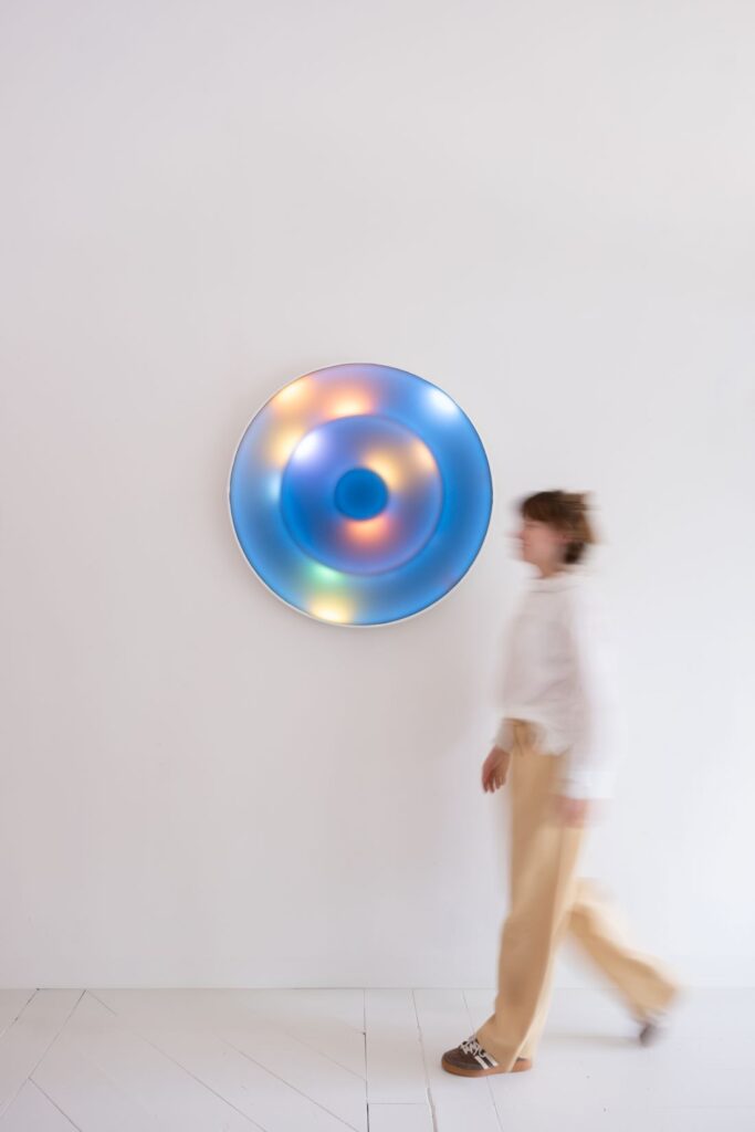A+N Studio designer walking past a circular wall-mounted light installation glowing in blue, green and warm iridescent tones