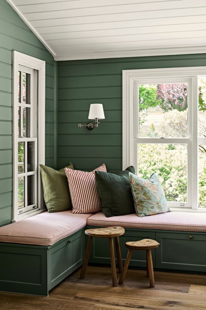 Close-up of a built-in green window seat with striped upholstery, layered cushions, timber stools and garden views