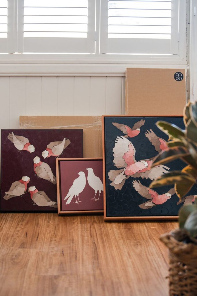 Three framed Australian bird artworks featuring stylised galahs and finches arranged on a timber floor in a sunlit home studio
