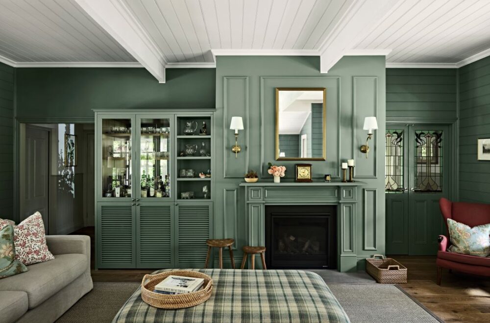 Formal sitting room with deep green panelled walls, a matching fireplace, glazed cabinetry, plaid ottoman and red armchair
