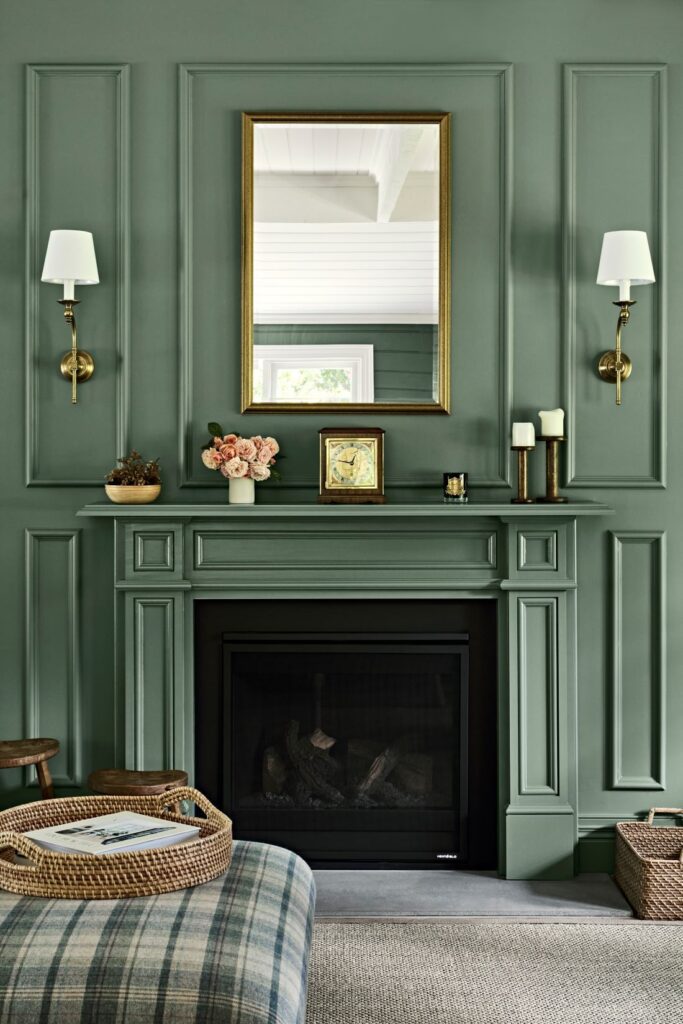 Deep green panelled fireplace wall with a gold-framed mirror, brass wall sconces and a plaid ottoman