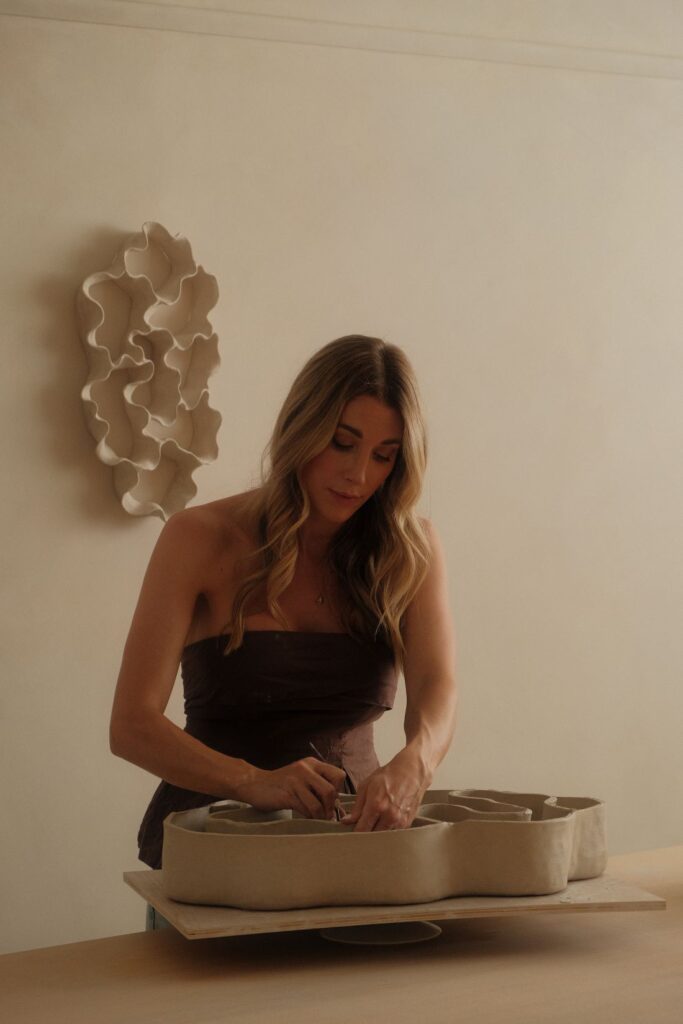 Ceramic artist Michelle Morton shaping a large hand-built clay form at a studio table, with an organic white wall sculpture behind her.