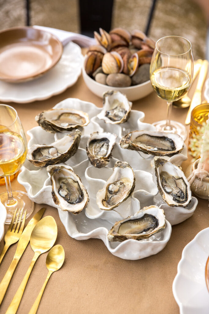 Fresh oysters arranged on a glossy white sculptural ceramic platter, surrounded by gold cutlery, glassware and layered tableware.