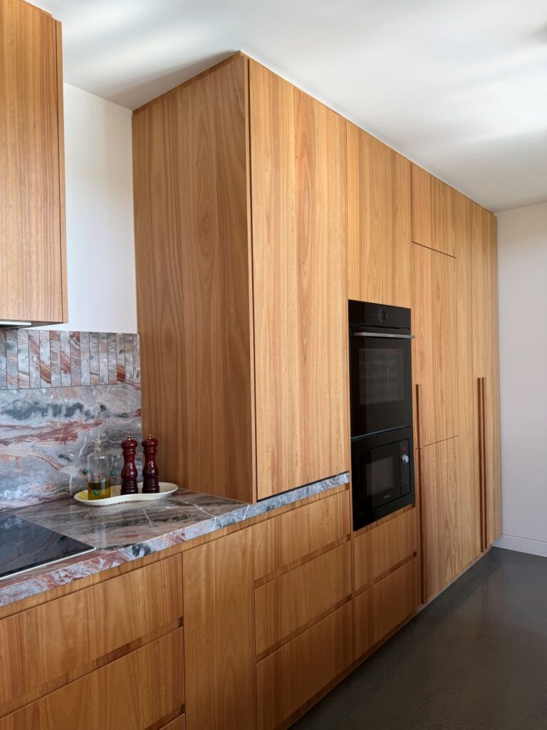 Warm timber kitchen cabinetry with integrated black appliances, stone benchtop, patterned stone splashback and styled kitchen accessories.