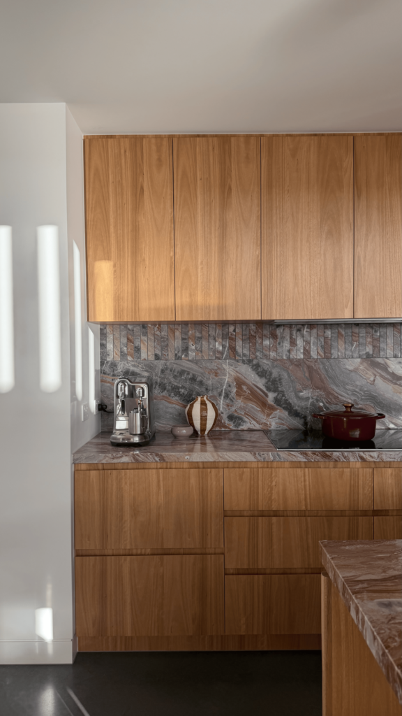 Warm timber kitchen cabinetry with richly veined stone benchtop and splashback, coffee machine, striped ceramic vase and burgundy cookware.