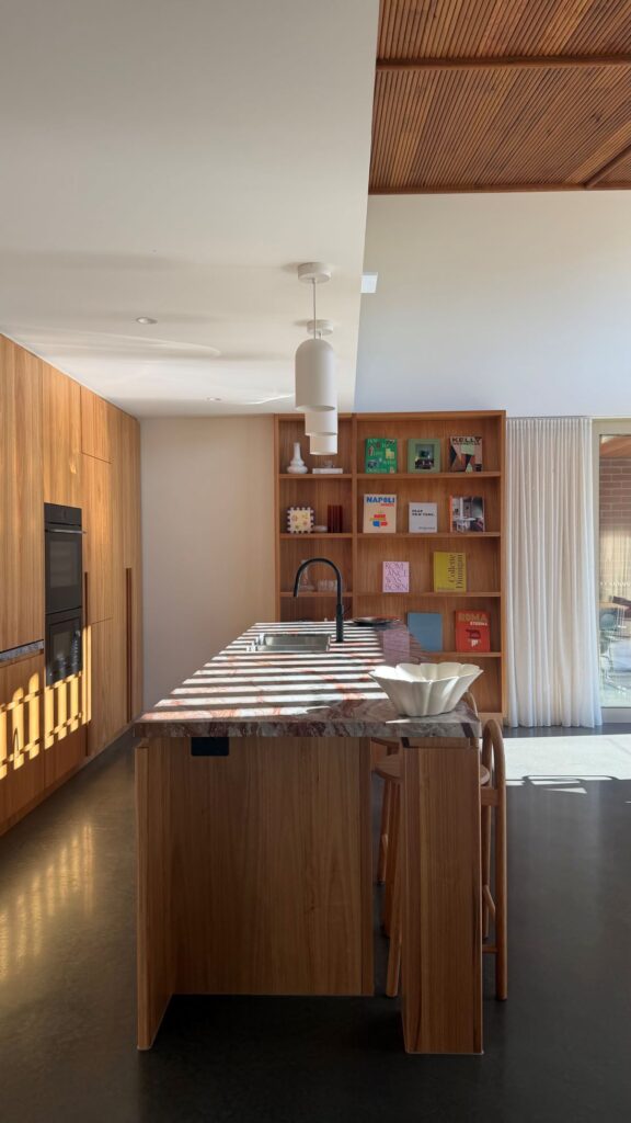 Timber kitchen island with dramatic stone benchtop, white pendant lights, black tapware and colourful art books on a built-in bookshelf.