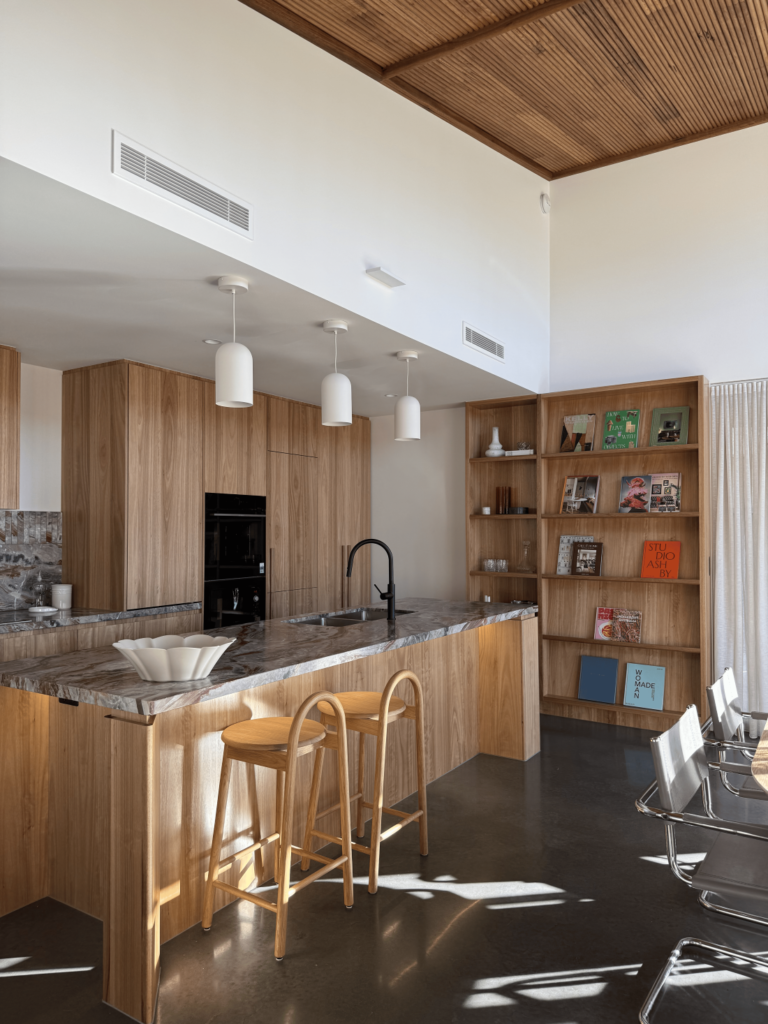 Light-filled timber kitchen with dramatic stone island, white pendant lights, woven bar stools, built-in bookshelf and timber batten ceiling.