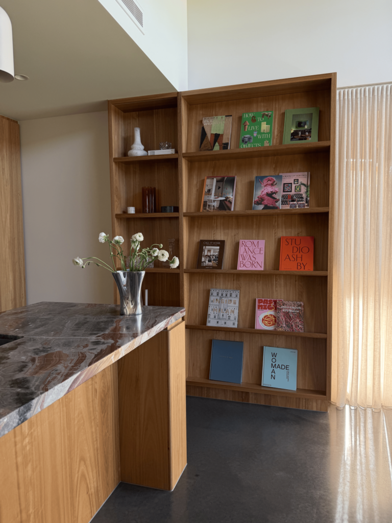 Timber kitchen island with dramatic stone benchtop beside a built-in bookshelf styled with art books, ceramics and soft sheer curtains.