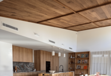 Inside a Canberra kitchen reno built around one incredible ceiling Open-plan kitchen, dining and living room with timber batten ceiling, warm wood cabinetry, cream sofa, patterned rug and sheer curtains.