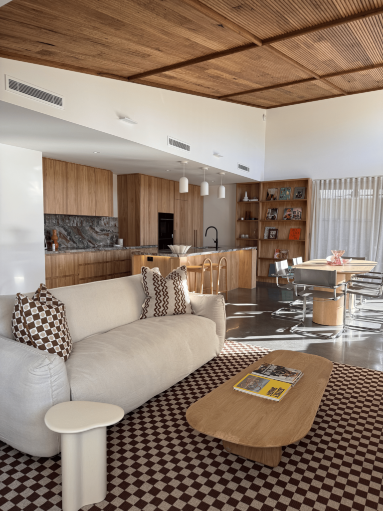 Open-plan kitchen, dining and living room with timber batten ceiling, warm wood cabinetry, cream sofa, patterned rug and sheer curtains.
