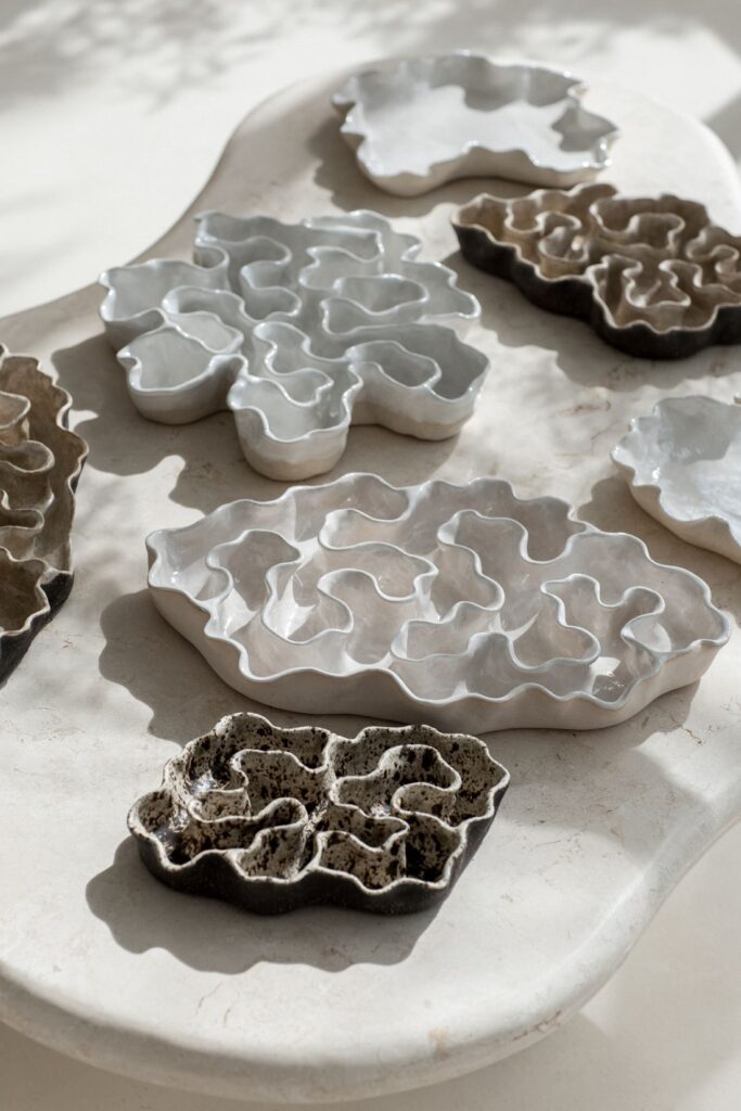 White, cream and dark glazed ceramic sculptures with layered wavy forms arranged on a pale stone table in natural sunlight.