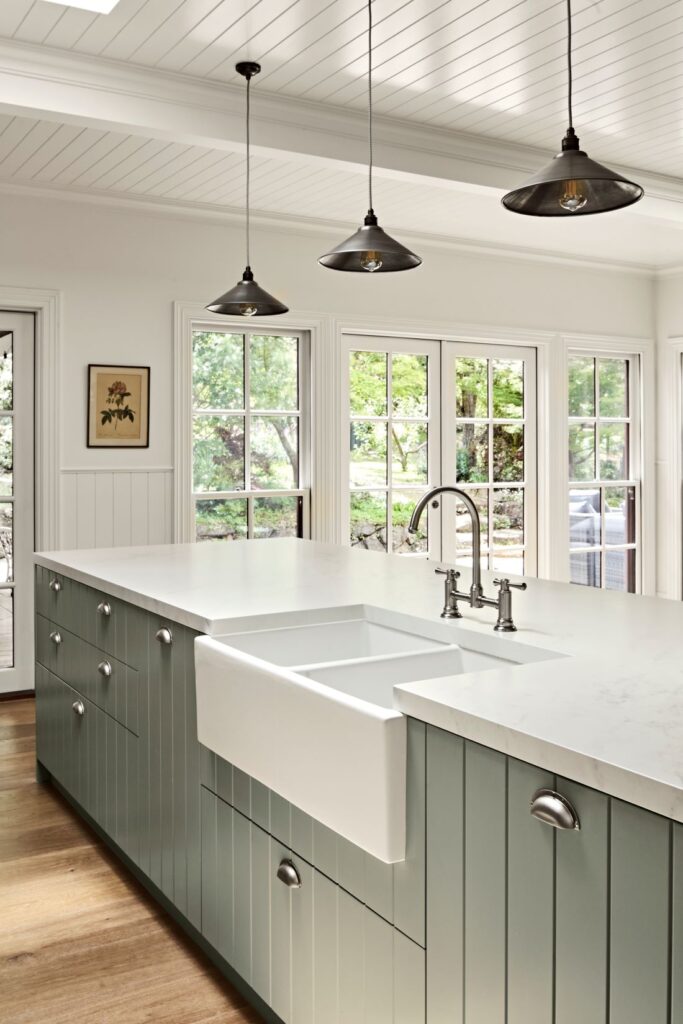 Sage green kitchen island with a white farmhouse sink, stone benchtop, chrome tapware and black pendant lights