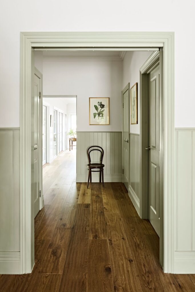 Light-filled hallway with sage green wall panelling, matching doors, warm timber flooring and framed botanical artwork