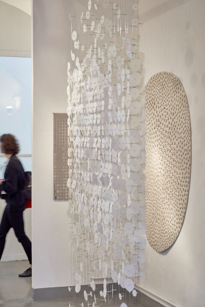 Hanging installation of white circular elements displayed beside an oval textured wall artwork in a gallery